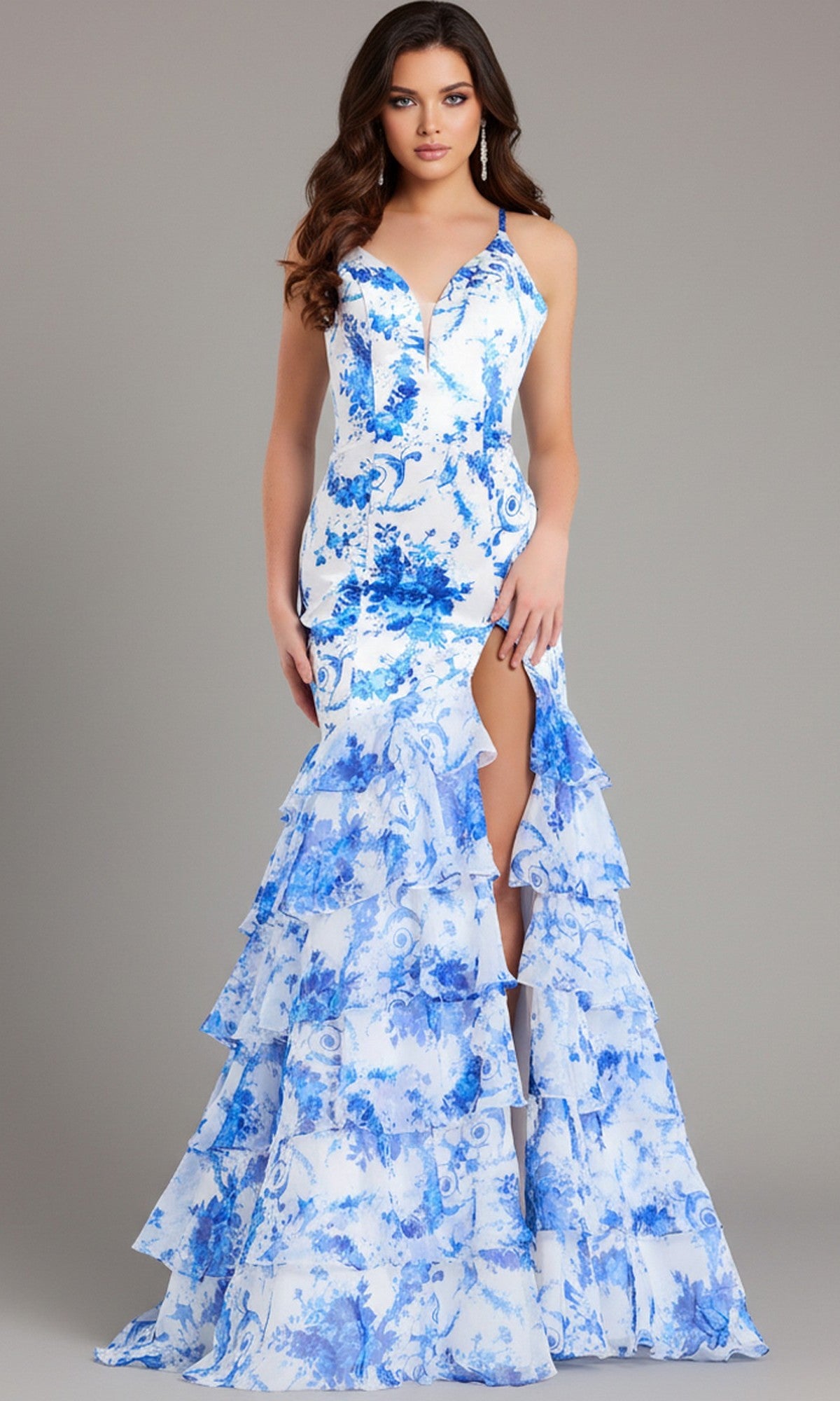 A woman models the Jovani 48331, a blue and white floral long prom dress with a tiered ruffle skirt, thigh-high slit, and lace-up open back, standing against a plain gray background.